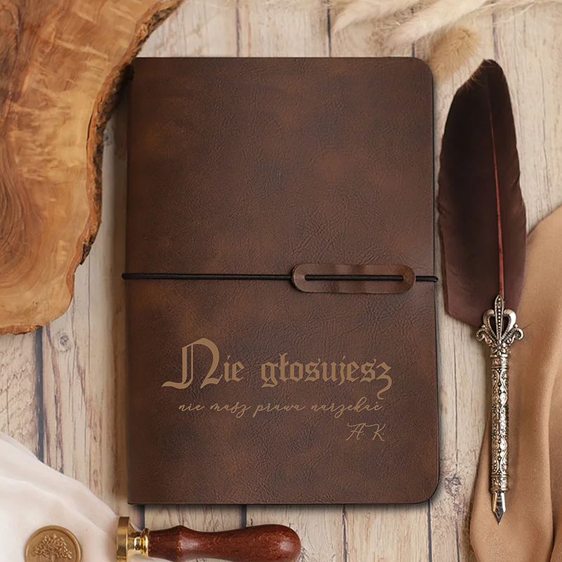 Brązowy notes o skórzanej okładce, z napisem 'Nie głosujesz' w ozdobnej czcionce, leżący na drewnianym blacie obok pióra gęsiego oraz metalowego stempla.