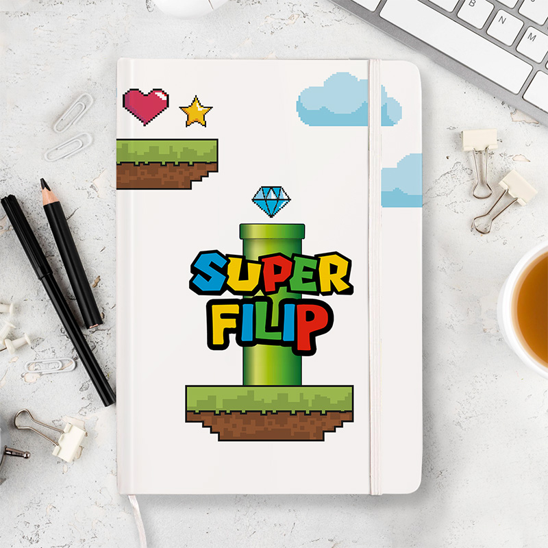 Notatnik z grafiką w stylu retro, przedstawiającą zieleń rurę, diament, serce i gwiazdę, z napisem 'SUPER FILIP' w kolorowej, pixelowanej czcionce, otoczony akcesoriami biurowymi i filiżanką kawy na jasnym tle.