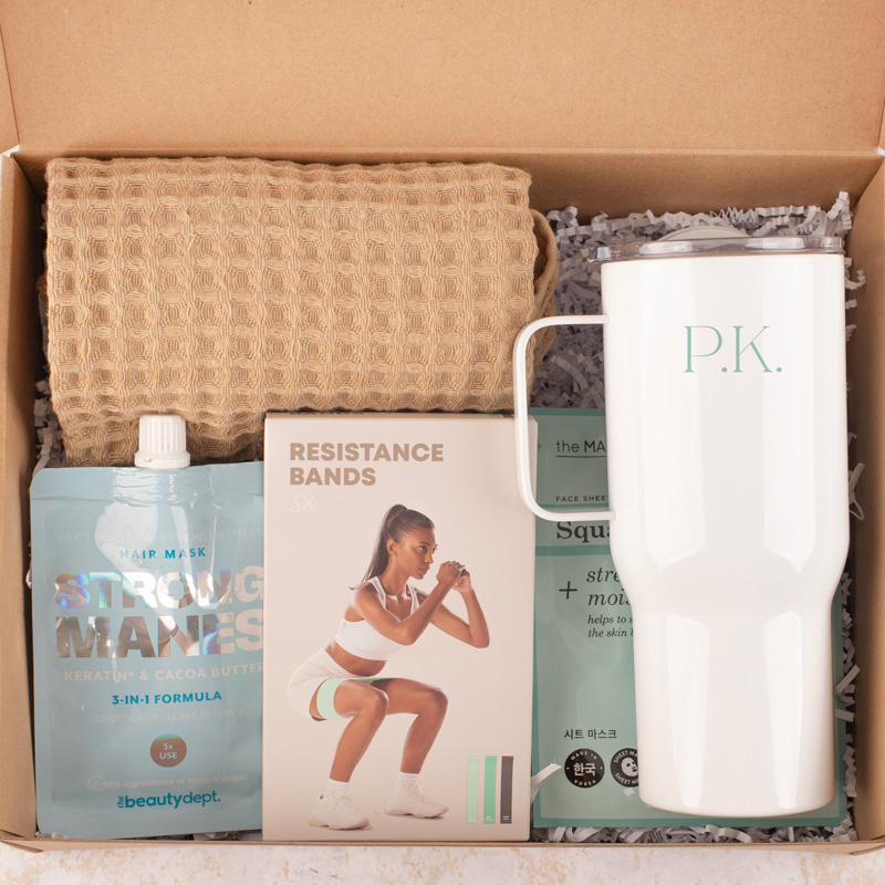 Zestaw prezentowy zawierający brązowy ręcznik, opakowanie ze wzmacniającą maską do włosów, instrukcję z oporowymi taśmami do ćwiczeń, maskę na twarz oraz białą termiczny kubek z inicjałami 'P.K.' w jasnym kolorze.