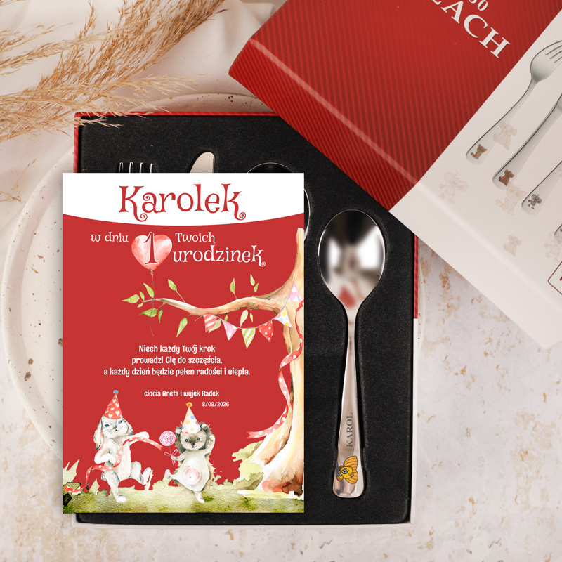 Urodzinowa kartka z imieniem "Karolek" oraz napisem "w dniu 1 Twoich urodzin" na czerwonym tle, otoczona ilustracjami dwóch wesołych zwierząt w kapeluszach urodzinowych, umieszczona na tle zestawu sztućców, w tym łyżki z grawerem, znajdującym się w eleganckim pudełku.