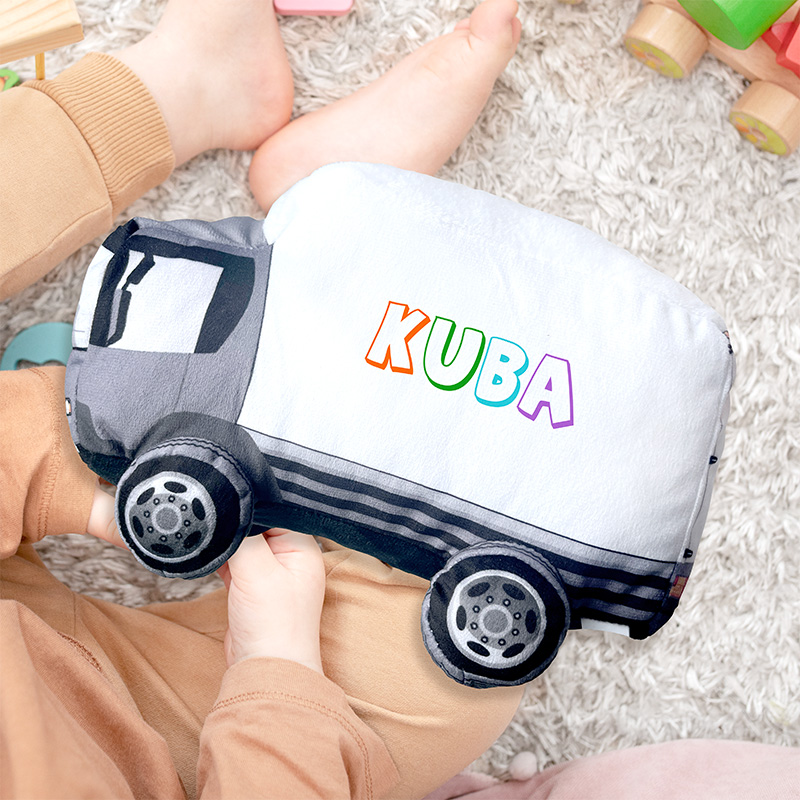 Dziecięce ręce trzymają miękką poduszkę w kształcie furgonetki z kolorowym napisem 'KUBA' na boku, leżące na dywaniku.
