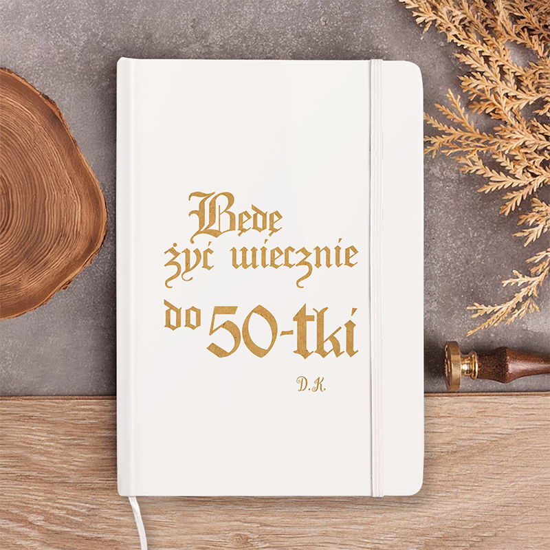 Biały notes z tekstem 'Będę żyć wiecznie do 50-tki' w złotym kolorze, obok drewniany krążek i stempel, w otoczeniu suchego kwiatu.