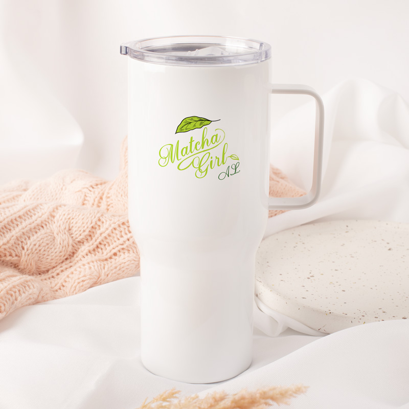 Biały kubek termiczny z nadrukiem "Matcha Girl" i zielonym liściem, stojący na miękkim różowym tle z dzianiny i jasnym tle z tkaniny.