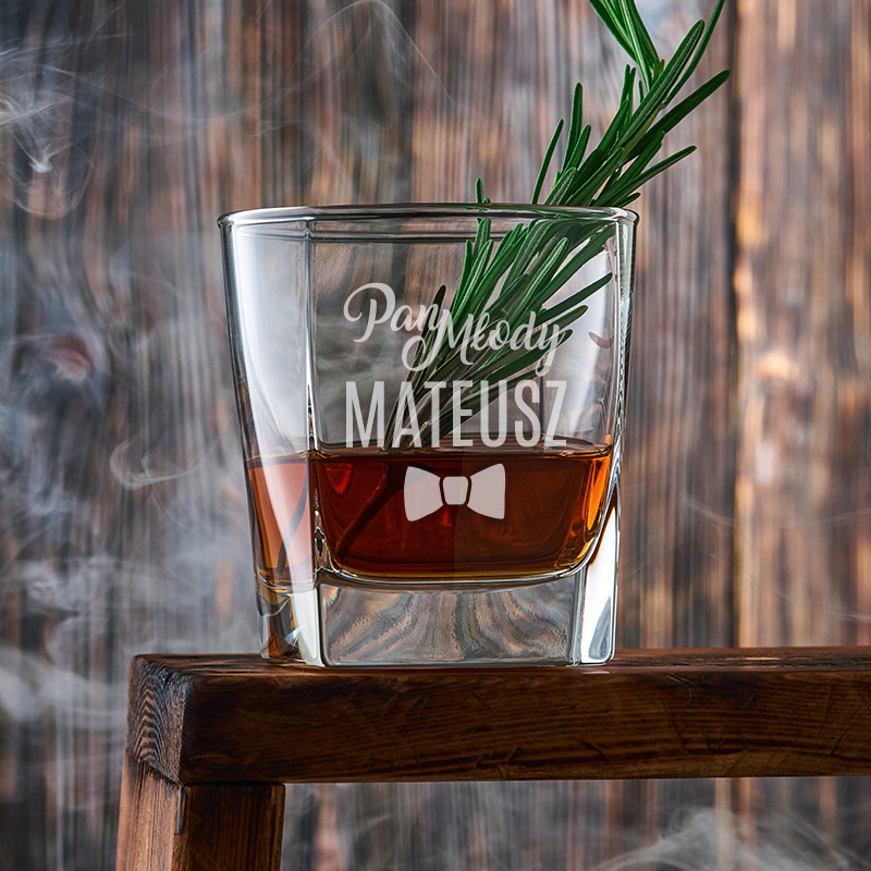 Szklanka z brązowym napojem, ozdobiona gałązką rozmarynu, z wygrawerowanym imieniem 'Mateusz' oraz napisami, stojąca na drewnianym blacie, w tle drewniana ściana i mgła.