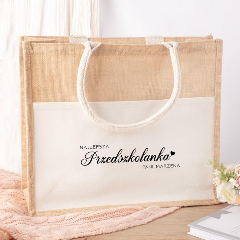 Torba materiałowa w stylu eko, z beżowym tłem i białym panelem, na którym znajduje się napis "NAJLEPSZA Przedszkolanka Pani Marzena" w eleganckiej czcionce, z plecionymi uchwytami.