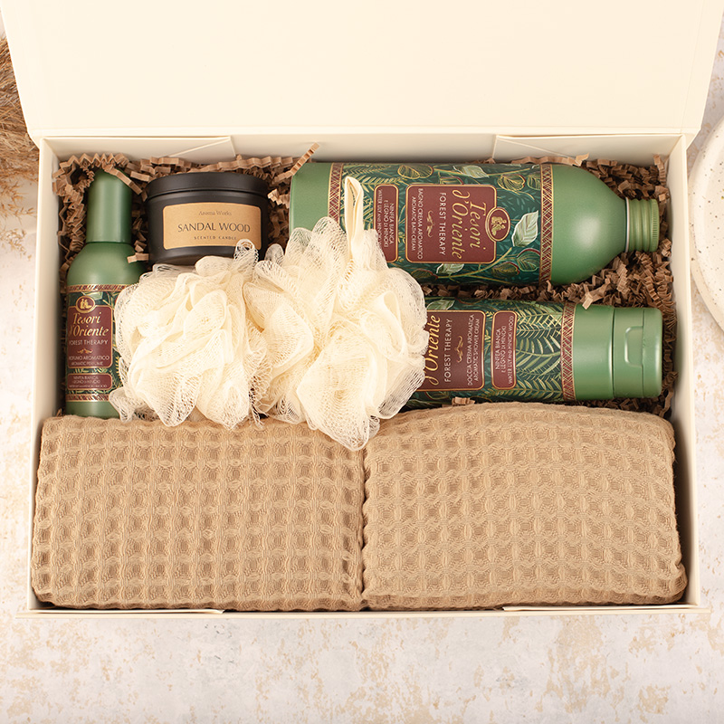 Zestaw produktów do pielęgnacji zawierający dwa szampony w zielonych butelkach, spray do włosów, opakowanie świecy zapachowej o zapachu sandałowca oraz dwa kremowe myjki w kształcie kwiatu, a także dwa beżowe ręczniki frotte w pudełku.