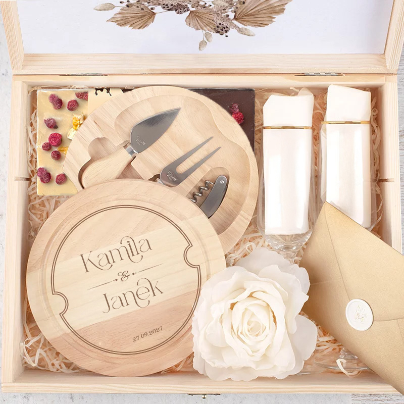 Elegancka skrzynka prezentowa z grawerowanymi deskami do serwowania, nożami oraz zestawem dwóch białych szklanek i złotą kopertą, obok sztucznej białej róży.