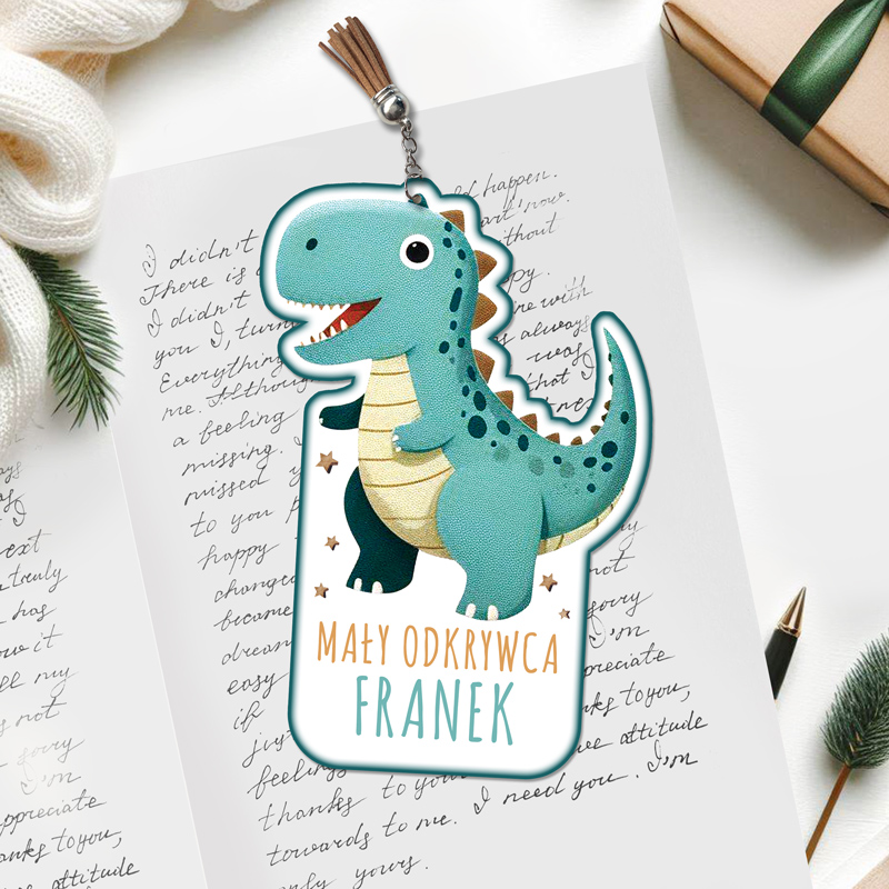 Ilustracja uroczej, turkusowej postaci dinozaura z uśmiechem, na tle notatek z ręcznym pismem i ozdobną paczką prezentową, z napisem 'MAŁY ODKRYWCA FRANEK' i pomponem na górze.