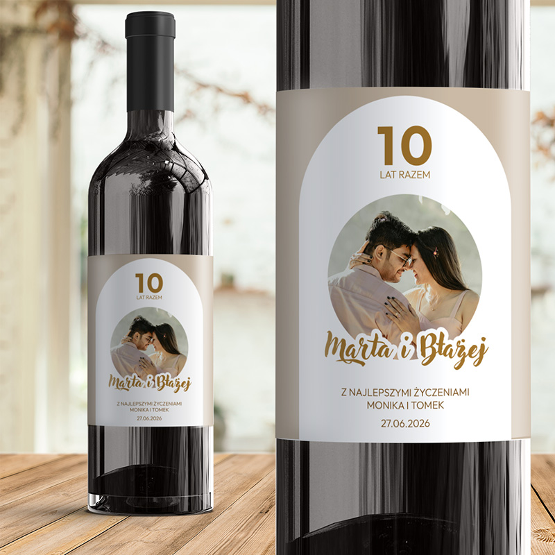 Butelka wina z etykietą na której widnieje serce, napis '10 lat razem', imiona 'Marta i Błażej', oraz życzenia od Moniki i Tomka z datą 27.06.2026, na tle drewnianego stołu i rozmytego tła w pomieszczeniu.