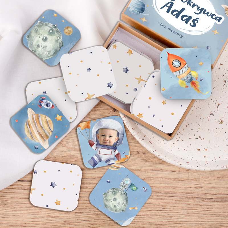 Gra memory składająca się z kolorowych kart z ilustracjami astronauty, rakiety, planet i kosmicznych obiektów, leżących na drewnianym blacie obok pudełka z napisem 'Odkrywca Adaś'.