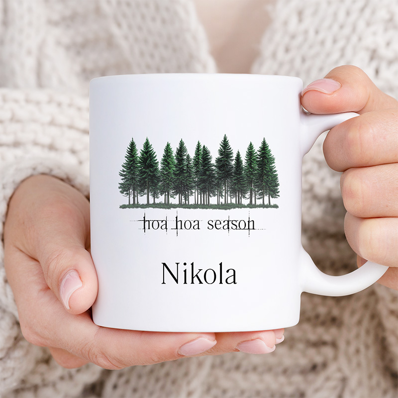 Biała filiżanka trzymana w dłoniach, z nadrukiem przedstawiającym rzędy zielonych drzew oraz napisem 'hoa hoa season' i imieniem. W tle widoczny jest jasny sweter.