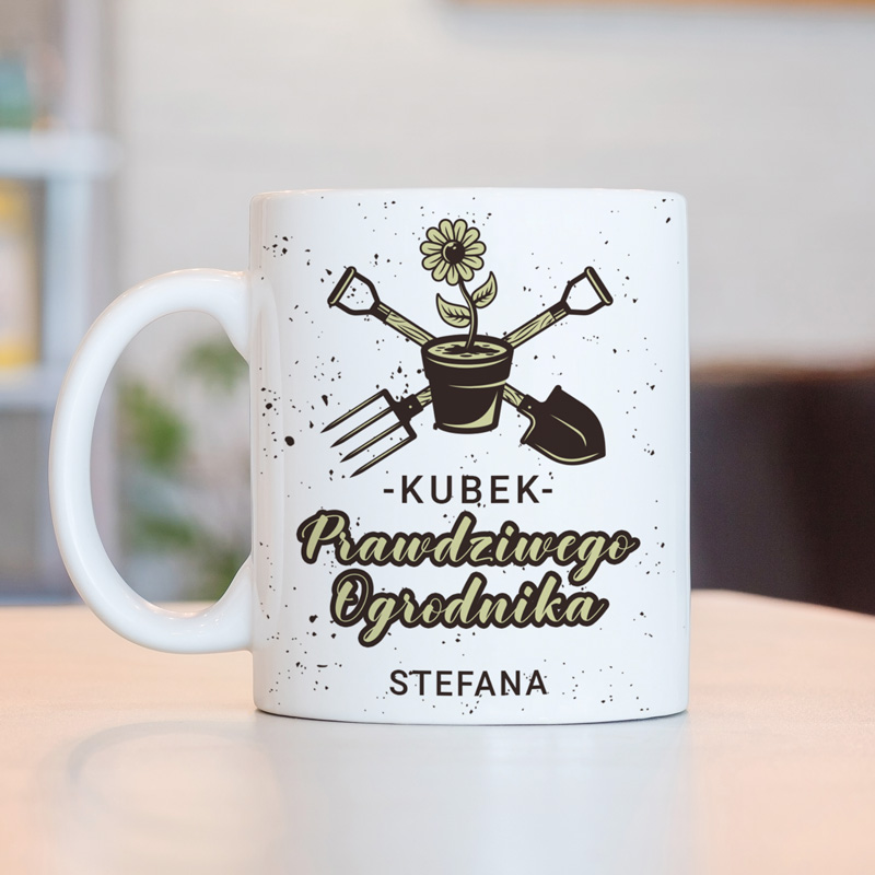 Kubek w kolorze białym z grafiką przedstawiającą doniczkę z kwiatem oraz narzędziami ogrodniczymi. Na kubku widnieje napis "KUBEK Prawdziwego Ogrodnika".
