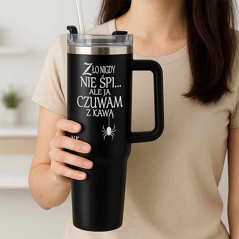Osoba trzyma czarny kubek termiczny z napisem 'Zło nigdy nie śpi... ale ja czuwam z kawą', z przezroczystą pokrywką i słomką, na tle minimalistycznego wnętrza.