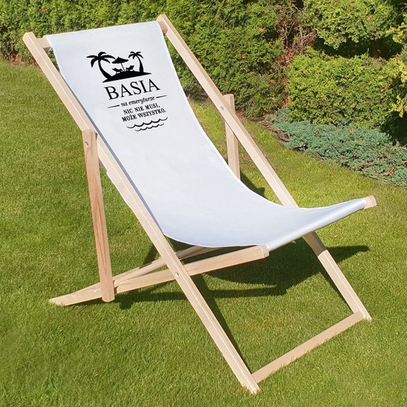 Leżak w białym kolorze z drewnianą ramą, z nadrukiem zawierającym napis 'BASIA' oraz tekstem w języku polskim, umieszczony na zielonej trawie w otoczeniu roślinności.