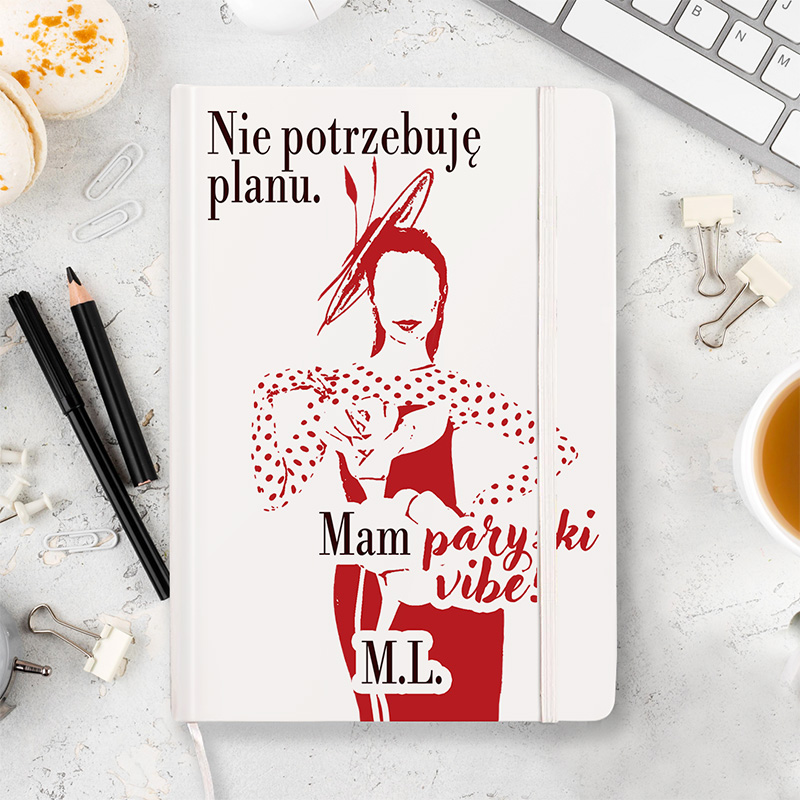 Notatnik z wizerunkiem kobiety w czerwonym ubraniu w grochy, trzymającej filiżankę. Na okładce napis: 'Nie potrzebuję planu. Mam paryski vibe! M.I.' W tle widoczne przybory biurowe i filiżanka kawy.