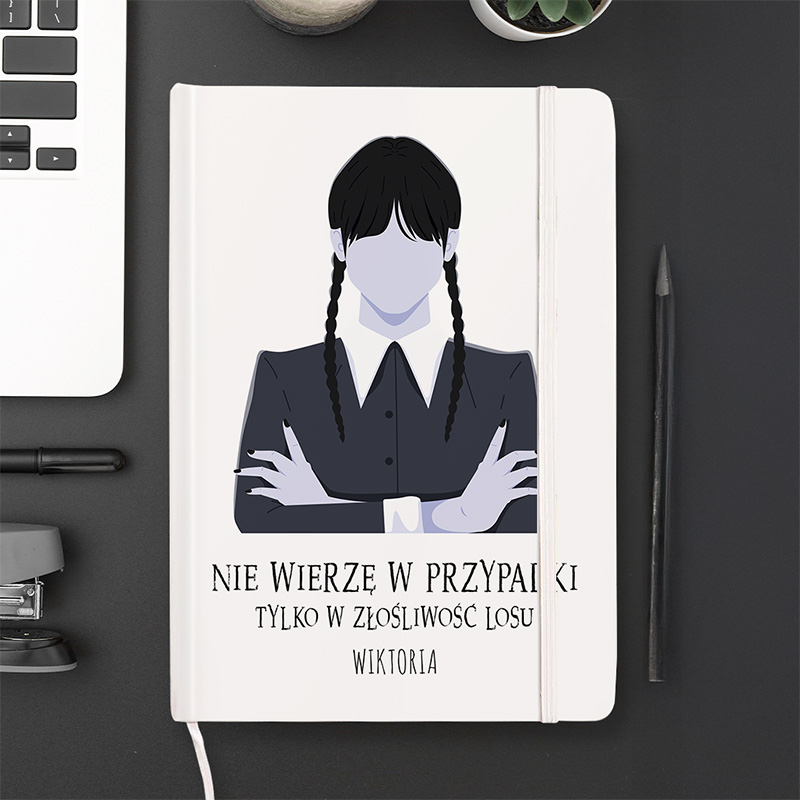 Nota książkowa z ilustracją postaci z długimi warkoczami, ubranej w czarną marynarkę, z podpisem "NIE WIERZĘ W PRZYPADKI TYLKO W ZŁOŚLIWOŚĆ LOSU WIKTORIA", umieszczona na ciemnym biurku obok laptopa, długopisu i rośliny doniczkowej.