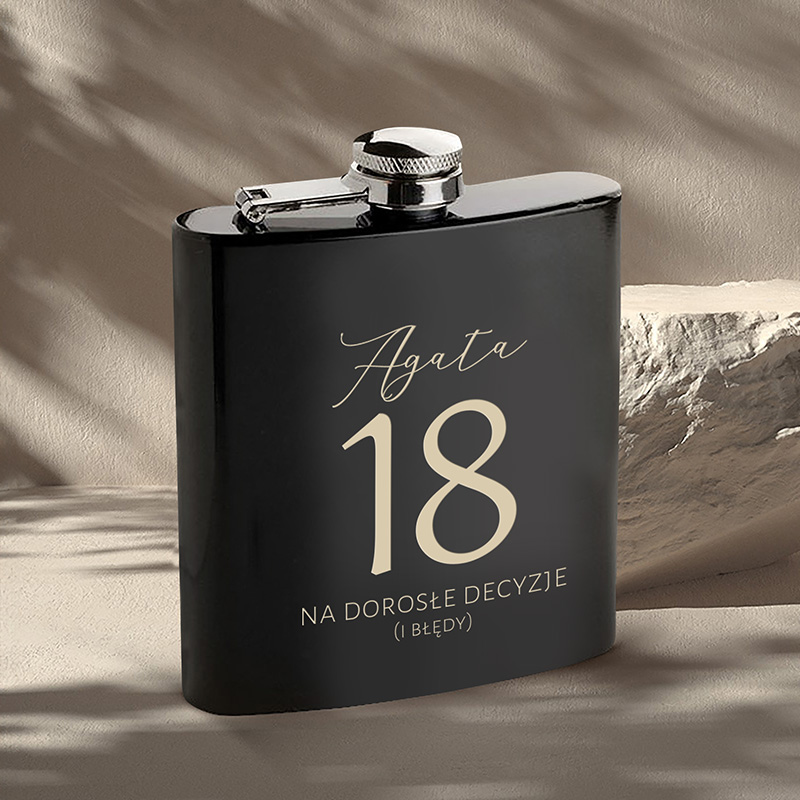 Czarna piersiówka z napisem 'Agata 18 NA DOROSŁE DECYZJE (I BŁĘDY)' w eleganckiej czcionce, umieszczona na tle jasnej powierzchni z delikatnym oświetleniem.