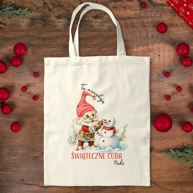 Torba płócienna z ilustracją gnomka w czerwonym kapeluszu, trzymającego marchewkę i stojącego obok bałwanka. Na torbie zapisano tekst "Tu mieszkają Świąteczne Cuda" oraz imię "Nadii", a w tle znajdują się czerwone ozdoby bożonarodzeniowe i gałązki choinki na drewnianym stole.