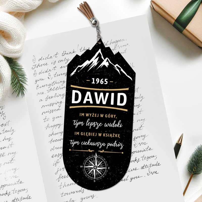 Zakładka do książki w kształcie góry z napisem 'DAWID' oraz rokiem '1965', zawierająca tekst w języku polskim i symbol kompasu, położona na kartce z zapisaną ręcznie notatką i ozdobnym tłem z gałązką świerku oraz prezentem zapakowanym w papier z kokardą.