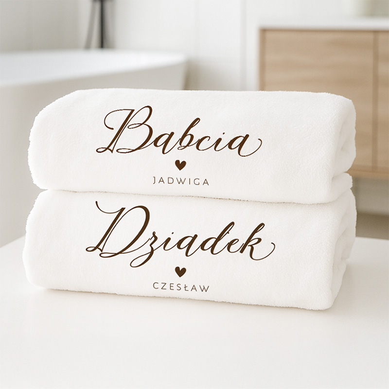 Dwa białe ręczniki leżące na jasnym stole, z nadrukowanymi imionami: na górnym ręczniku "Babcia Jadwiga" i na dolnym "Dziadek Czestław", oddzielone sercem.
