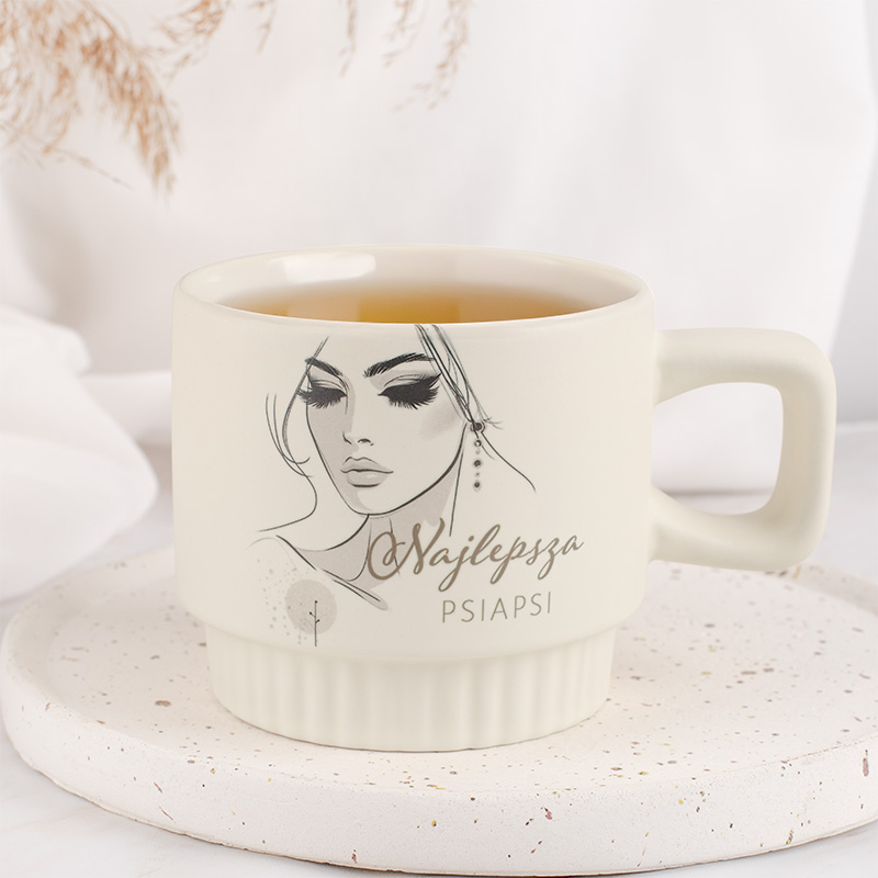 Jasna filiżanka z rysunkiem kobiety i napisem "Najlepsza Psiapsi" na białym talerzyku, otoczona jasnym tłem oraz delikatnymi elementami dekoracyjnymi.