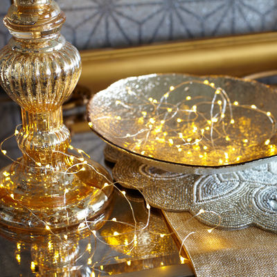 Szklany świecznik z bombkami świetlnymi obok złotej tacy i haftowanego obrusu w eleganckim wnętrzu.