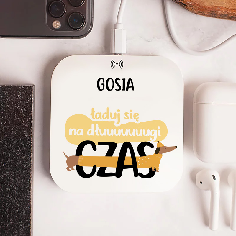 Design of a wireless charger with the text "ładuj się na dłuuuuuuuugi CZAS" and a cartoon illustration of a dachshund. The charger is placed near a smartphone and white earbuds on a light surface.