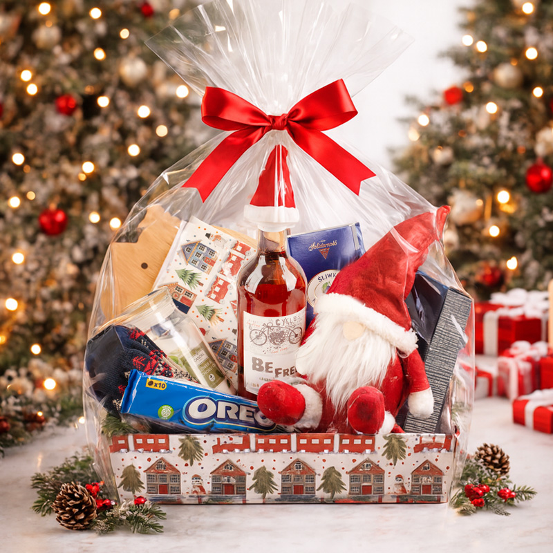 Kosz prezentowy z przezroczystą folią, ozdobiony czerwoną wstążką. Zawiera pluszowego Mikołaja, butelkę napoju, opakowanie ciasteczek Oreo, a także różne przedmioty świąteczne oraz słodycze. W tle widać rozświetlone choinki i choinkowe dekoracje, co tworzy bożonarodzeniową atmosferę.