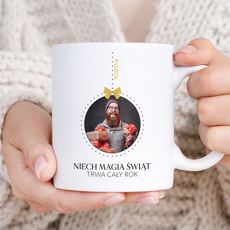 Biała filiżanka trzymana przez osobę w swetrze, z napisem "NIECH MAGIA ŚWIĄT TRWA CAŁY ROK" oraz okrągłym zdjęciem mężczyzny z brodą w okularach, trzymającego prezenty z czerwonymi kokardami, na przodzie filiżanki.