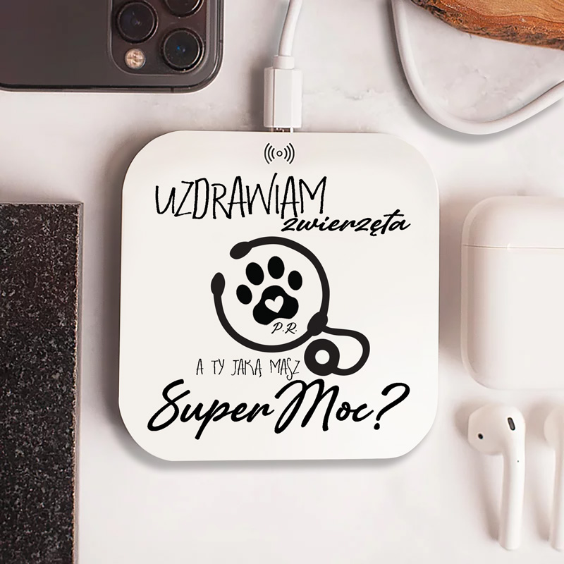 Podstawka do ładowania z napisem "UZDRAWIAM zwierzęta" i ilustracją łapki z sercem oraz tekstem "a Ty jaką masz Super Moc?". W tle widoczny telefon i słuchawki bezprzewodowe.