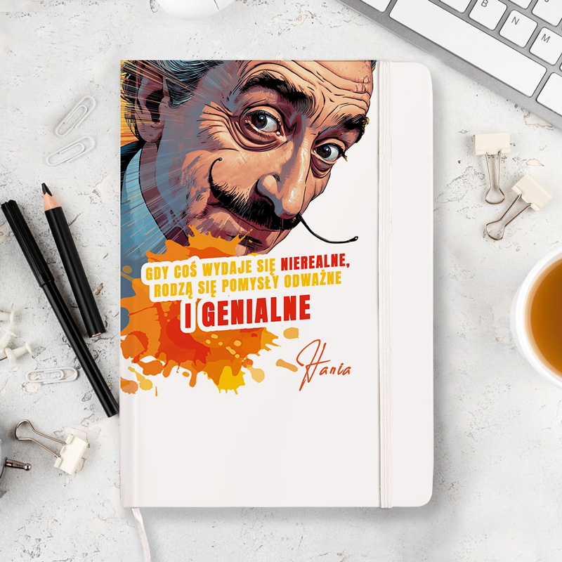 Kreatywny notes z ilustracją mężczyzny w związku z cytatem: 'Gdy coś wydaje się nierealne, rodzą się pomysły odważne i genialne'. W tle kolorowe plamy farby, na biurku obok długopisy oraz filiżanka z kawą.