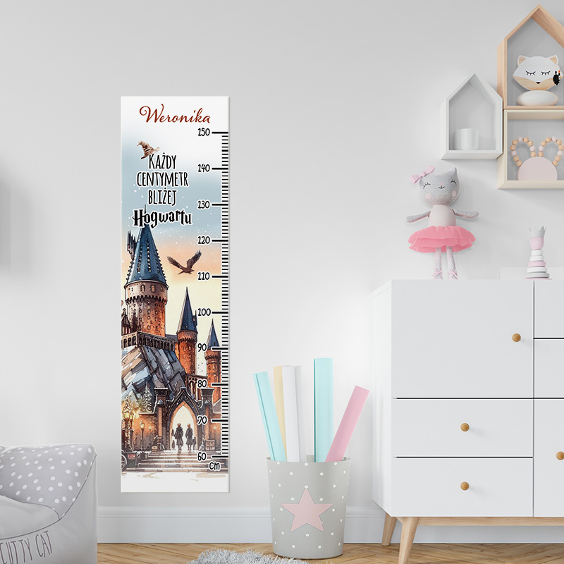 Drabina wzrostu, z ilustracją zamku przypominającego Hogwart, z miarką w centymetrach, podzielona na różne wysokości, z napisem 'Każdy centymetr bliżej Hogwartu' oraz imieniem 'Weronika', umieszczona na jasnej ścianie obok kolorowych kartek w pojemniku.