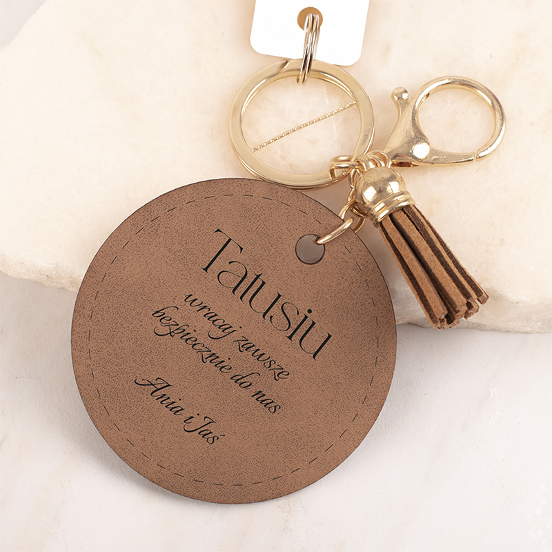 Okrągły breloczek w kolorze brązowym z tekstem 'Tatusiu' i napisem informującym o bezpieczeństwie. Dołączony złoty kluczyk z frędzlami.
