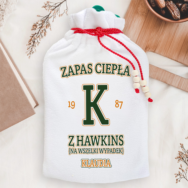 Biała torba z zasuwaniem, z nadrukiem w stylu retro, zawierającym napis 'ZAPAS CIEPŁA', dużą literę 'K' oraz rok '1987', a na dole napis 'Z HAWKINS (NA WSZELKI WYPADEK) KLAUDIA'. Torba ozdobiona czerwoną sznurówką i drewnianymi koralikami, leżąca na jasnym tle z dekoracjami.