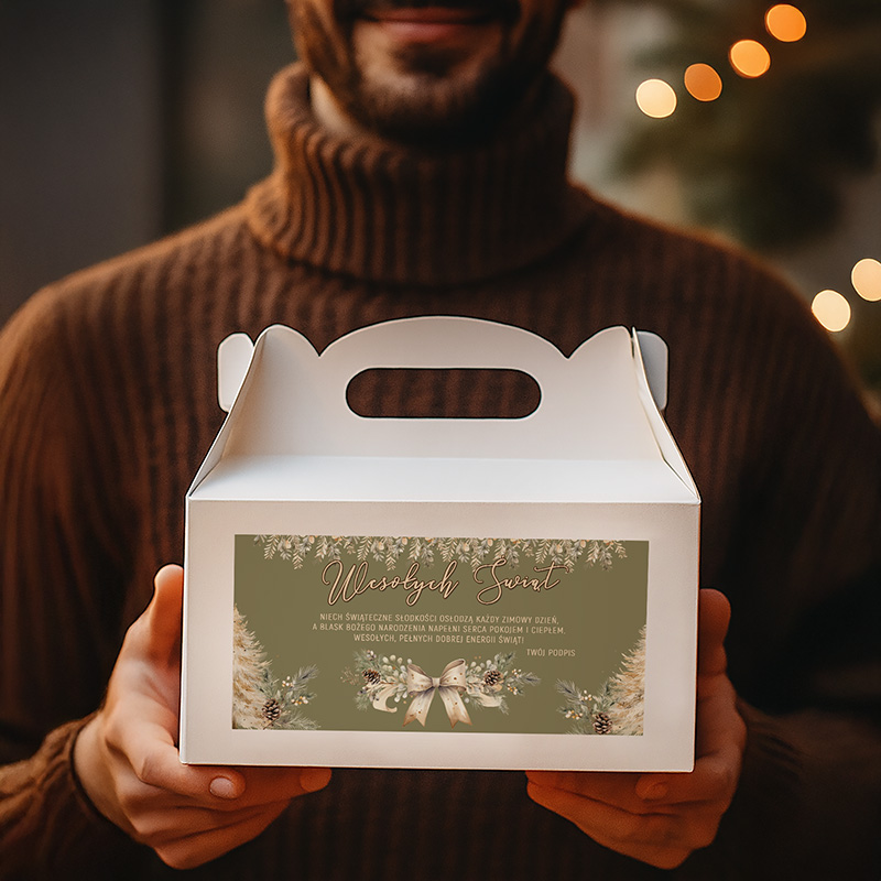 Mężczyzna w brązowym swetrze trzyma białe pudełko z uchwytem. Na pudełku znajduje się świąteczny napis 'Wesołych Świąt' oraz ozdobne ilustracje roślinne z wstążką. W tle widać rozmyte świąteczne lampki.