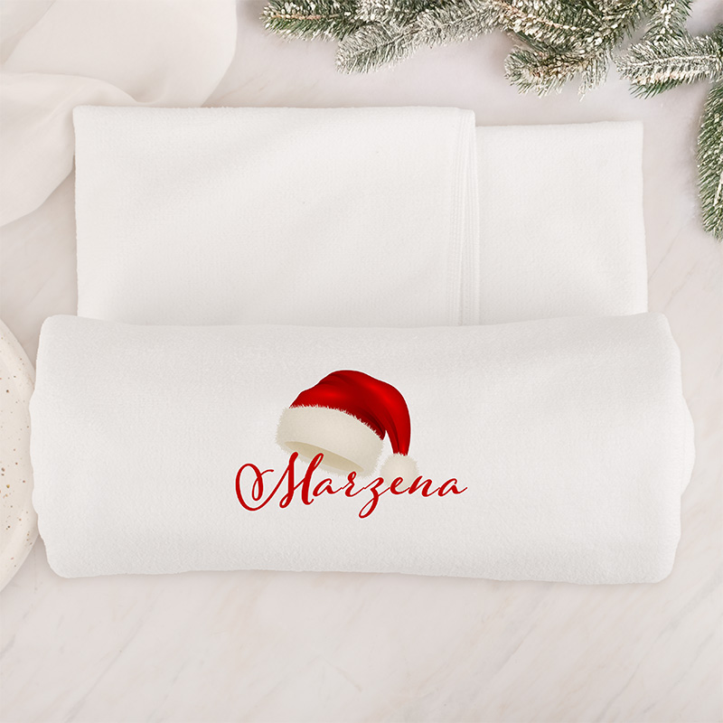 Biała narzuta z czerwonym napisem "Marzena" i ilustracją czerwonej czapki z białym pomponem, leżąca na jasnym tle z zielonymi gałązkami w tle.