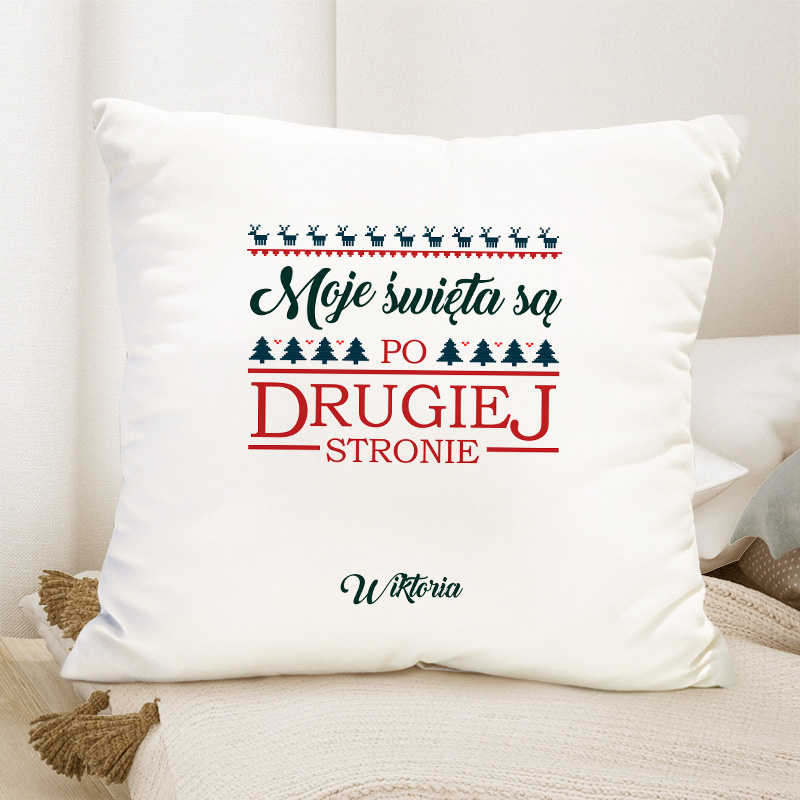 Poduszka z napisem „Moje święta są po drugiej stronie” w kolorze zielonym i czerwonym, udekorowana małymi choinkami i reniferami, umieszczona na tle jasnej narzuty oraz poduszki.