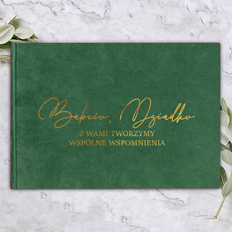 Zielona okładka książki z złotym napisem: "Babciu, Dziadku Z Wami Tworzymy Wspólne Wspomnienia". Tło w marmurowym wzorze, a po prawej stronie liście roślinne.