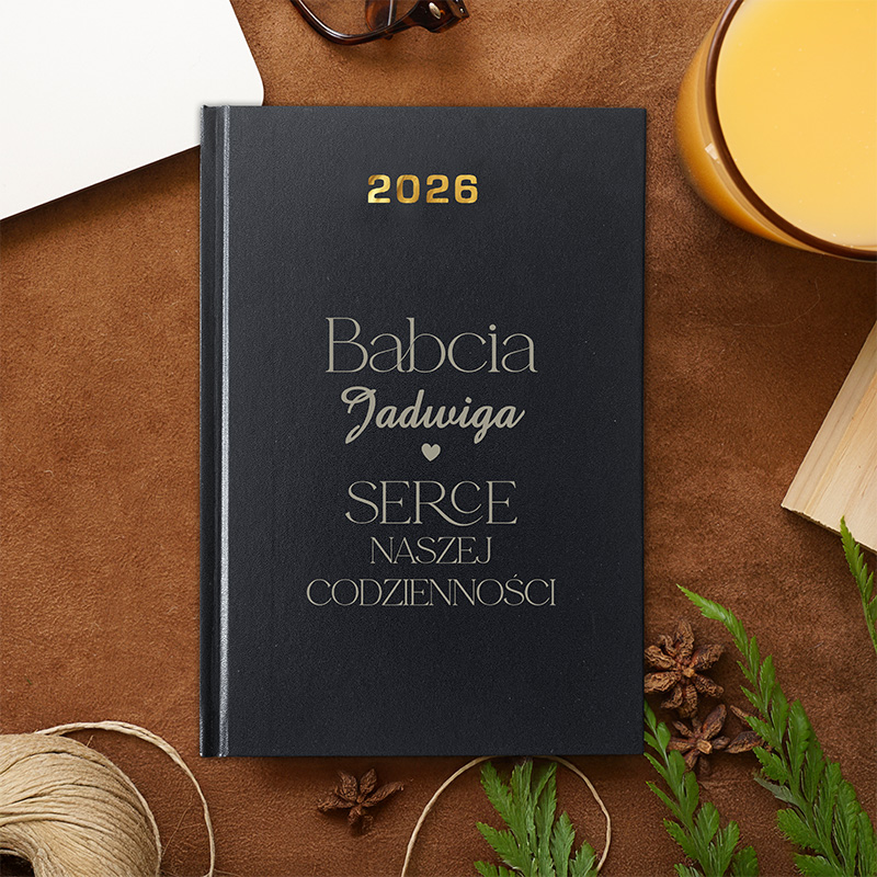 Czarna okładka książki z napisem "Babcia Jadwiga SERCE NASZEJ CODZIENNOŚCI" oraz rokiem "2026". W tle widoczne są okulary, szklanka z napojem oraz elementy dekoracyjne, takie jak gałązki i kawałek sznura.