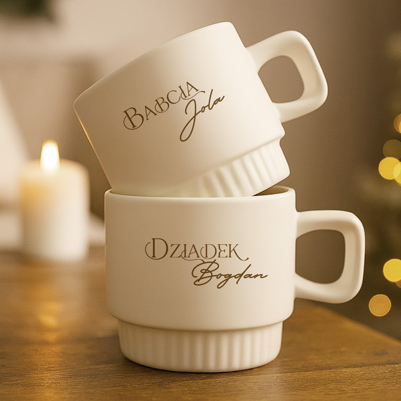 Na drewnianym stole znajdują się dwa białe kubki, jeden na drugim. Górny kubek ma napis 'Babcia', a dolny 'Dziadek'. W tle widoczna jest rozmyta świeca i elementy dekoracyjne ze światłami.