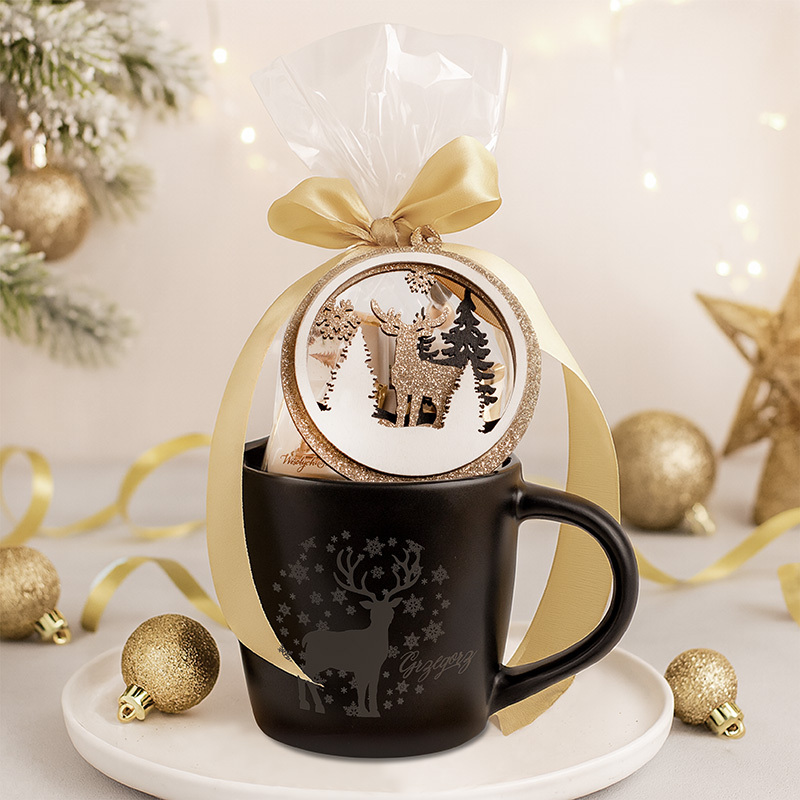 Czarny kubek z motywem renifera, ozdobiony złotą wstążką i bombką świąteczną, stoi na talerzyku obok złotych ozdób.