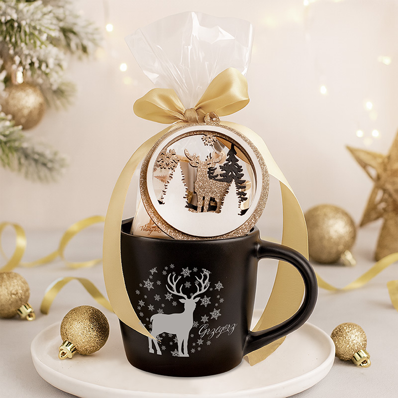 Czarna filiżanka z białym nadrukiem renifera i śnieżynkami, z ozdobnym opakowaniem w przezroczystą folię i złotą wstążką, a także dekoracyjnym medalionem z motywem leśnym; w tle świąteczne ozdoby na jasnośląskim stole.