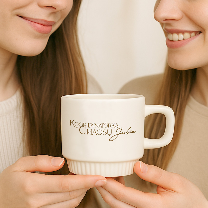 Dwie osoby trzymają ceramiczny kubek z napisem 'Koordynatorka Chaosu Julia', uśmiechając się do siebie.