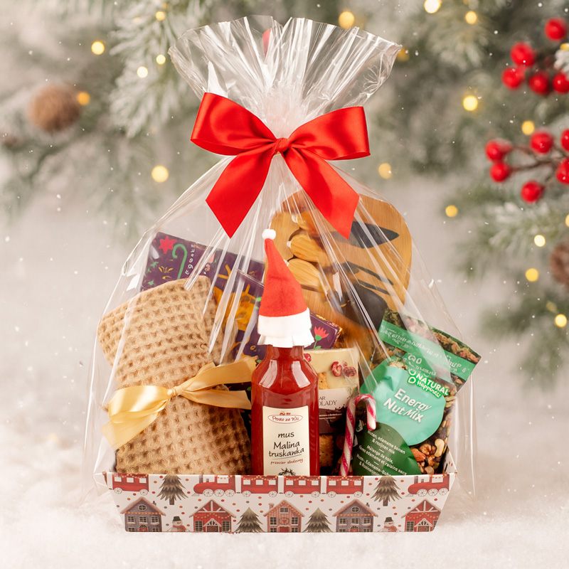 Kosz prezentowy ozdobiony przezroczystą folią i czerwoną kokardą, zawierający różnorodne smakołyki, takie jak dżem, przekąski i słodycze, w scenerii z choinką w tle i padającym śniegiem.