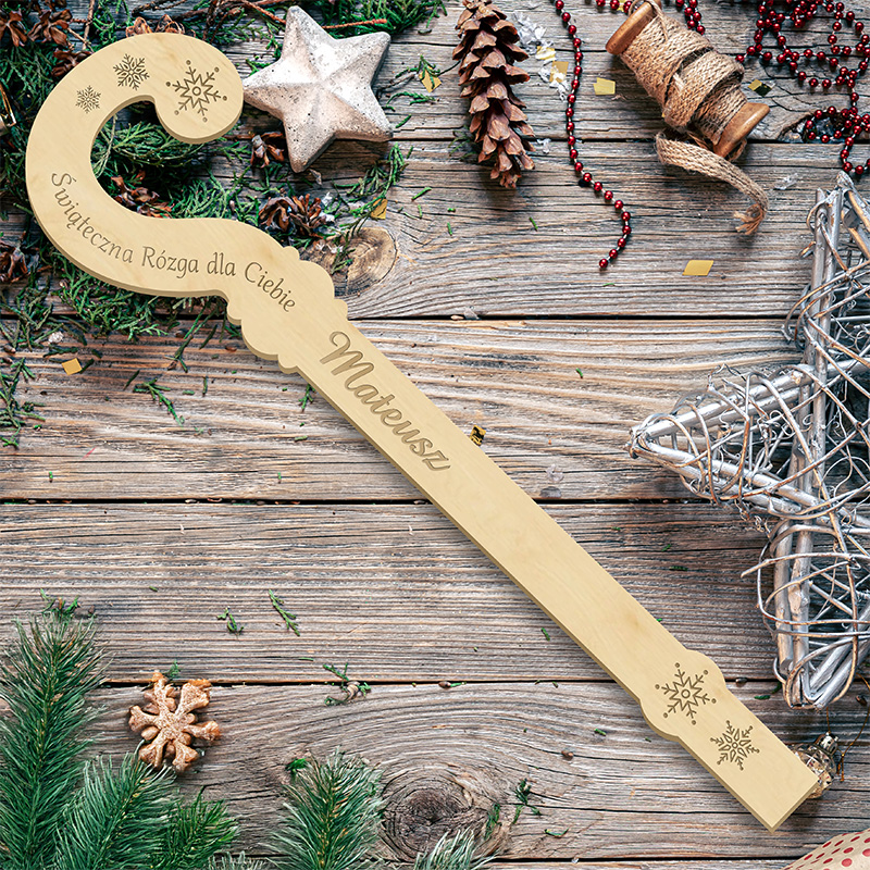 Drewniana, świąteczna rózga z napisem "Świąteczna Rózga dla Ciebie IMIĘ" leżąca na drewnianym stole, otoczona zielonymi gałązkami, szyszkami, świątecznymi dekoracjami i konfetti.