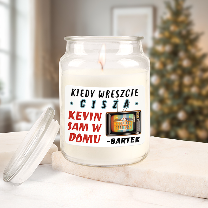 Świeca zapachowa w szklanym pojemniku z etykietą, na której znajduje się napis 'KIEDY WRESZCIE CISZA KEVIN SAM W DOMU - IMIĘ', w tle widać rozmyte drzewko świąteczne i przytulne wnętrze.