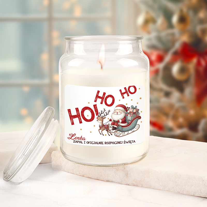 Świeca w szklanym słoiku z etykietą przedstawiającą Świętego Mikołaja na saniach ciągniętych przez renifera, z napisem "HO HO HO" oraz polskim tekstem "Zapal i oficjalnie rozpocznij święta". Słoik stoi na marmurowej powierzchni, w tle rozmyte dekoracje świąteczne.
