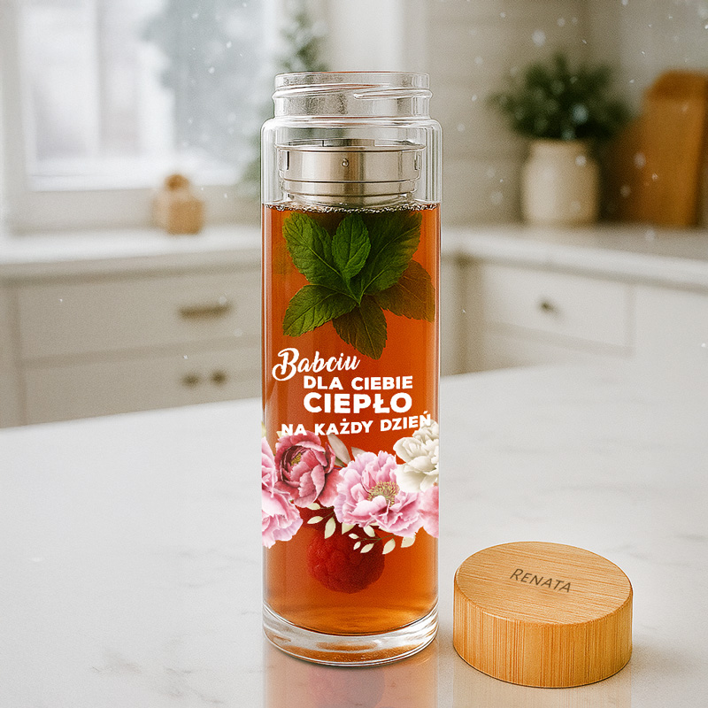 Przezroczysty pojemnik z herbatą z miętą i maliną, z napisem "Babciu DLA CIEBIE CIEPŁO NA KAŻDY DZIEŃ" oraz różowymi kwiatami u dołu, na tle jasnej kuchni z drewnianymi detalami.