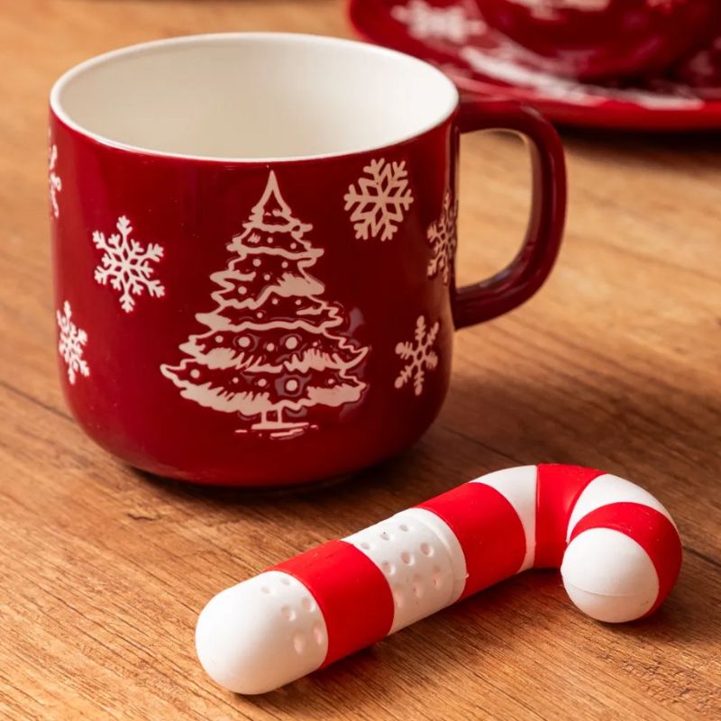 Czerwona filiżanka ozdobiona białym wzorem w kształcie choinki i płatków śniegu, obok niej znajduje się cukierek w paski czerwono-białe, na drewnianym blacie.