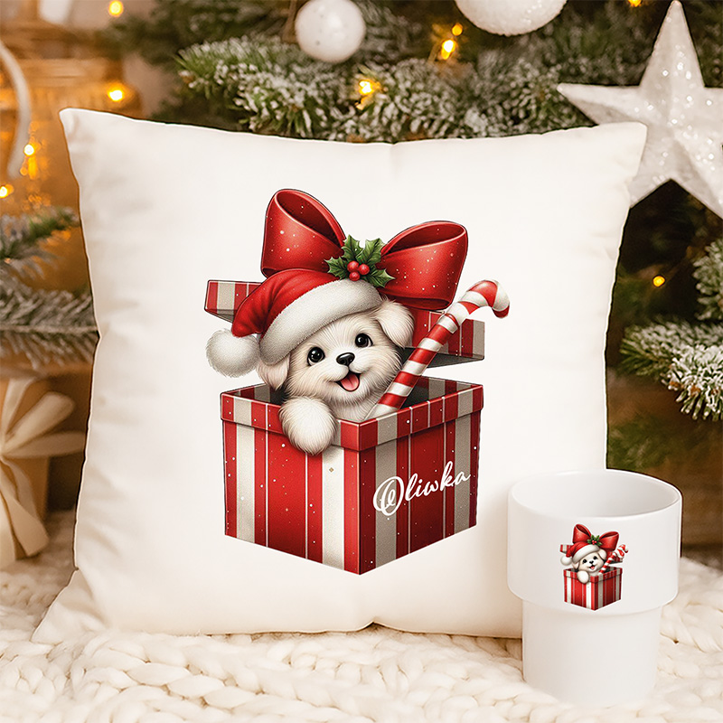 Poduszka z rysunkiem szczeniaka w czerwonym pudełku, noszącego świąteczny kapelusz i trzymającego laskę cukrową, obok kubek z tym samym motywem, w tle świąteczne dekoracje.